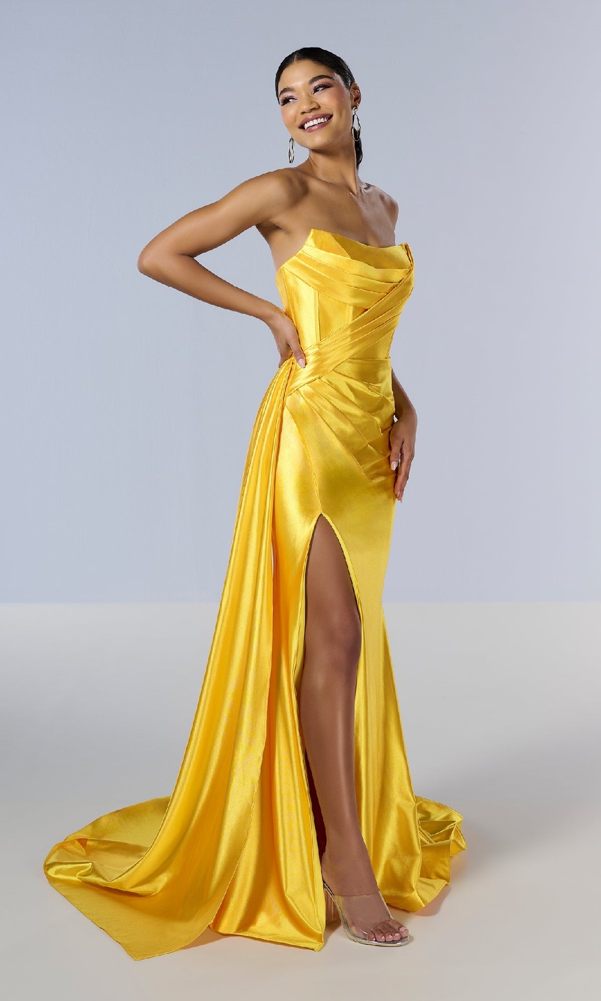 A woman poses and smiles against a plain light background, wearing the Tiffany 16178 long prom dress in bright yellow satin with a strapless corset bodice, thigh-high slit, and long train.